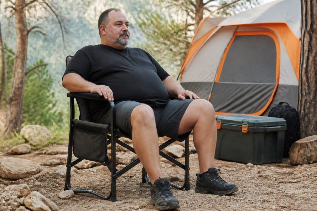 A heavy-set camper sitting comfortably in a durable steel-frame camping chair at a campsite, with a tent and outdoor gear visible in the background, showing the chair in active use on uneven terrain
