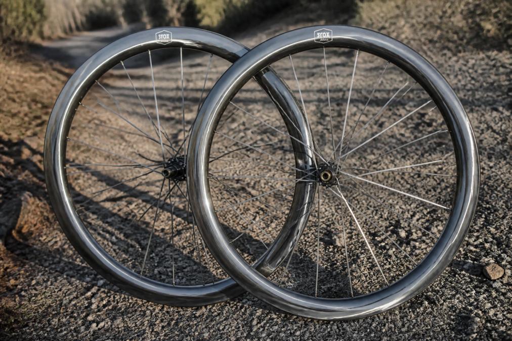 Close-up of a hand-built carbon gravel wheelset leaning against a gravel trail, showing visible carbon spoke detail and rim depth markings, with natural afternoon light casting shadows across the rim profile