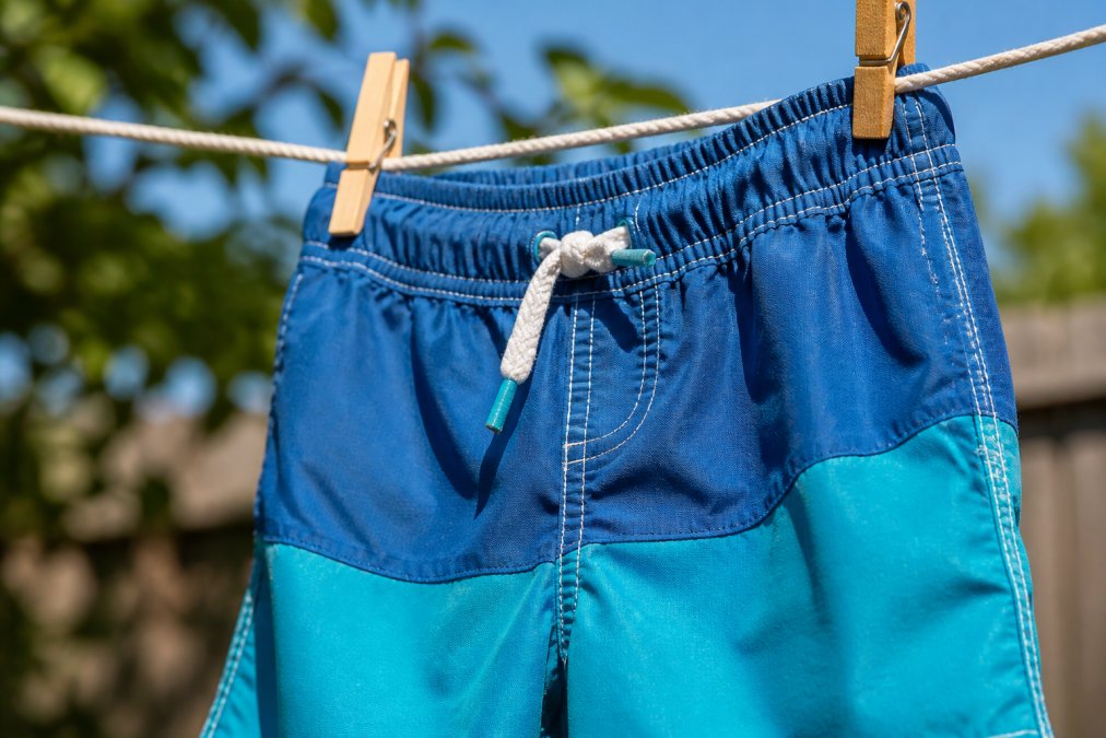 Close-up of a child's swim trunks hanging on a clothesline outdoors, showing fabric texture, reinforced seams, and elastic waistband in natural sunlight