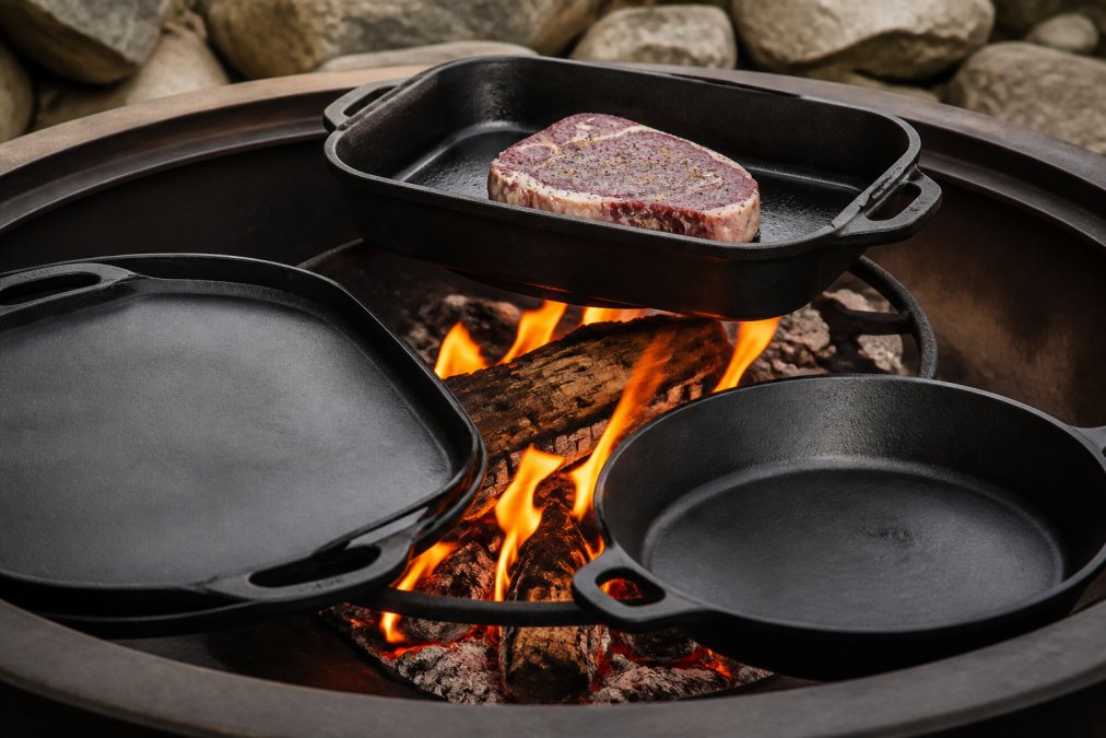 Close-up of cast iron cookware arranged around an active outdoor fire pit, showing a roasting pan, griddle, and skillet with flames in the background, golden firelight illuminating the cooking surfaces