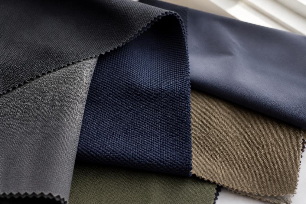 Close-up of different fabric swatches showing polyester blend textures, poly-cotton pique weave, and nylon elastane composites arranged under natural window lighting on a work surface