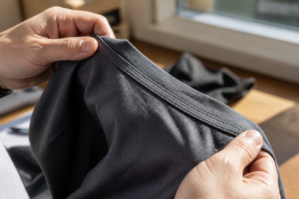 Close-up of a person's hands holding the collar and hem of a quality cotton t-shirt, pinching the double-needle stitching to show the tight, even seam detail in bright, natural window light