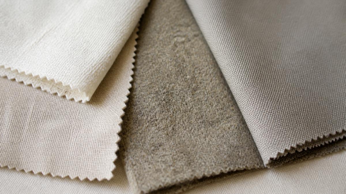 Close-up of fabric swatches showing texture differences between natural cotton, linen, wool, and synthetic materials laid out on a neutral surface with natural lighting