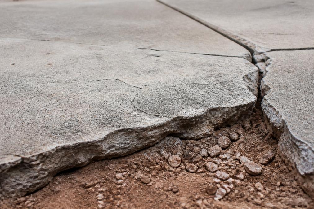Close-up of cracked concrete driveway surface showing visible separation and foundation settling, with exposed clay soil visible at the edges where material has shifted