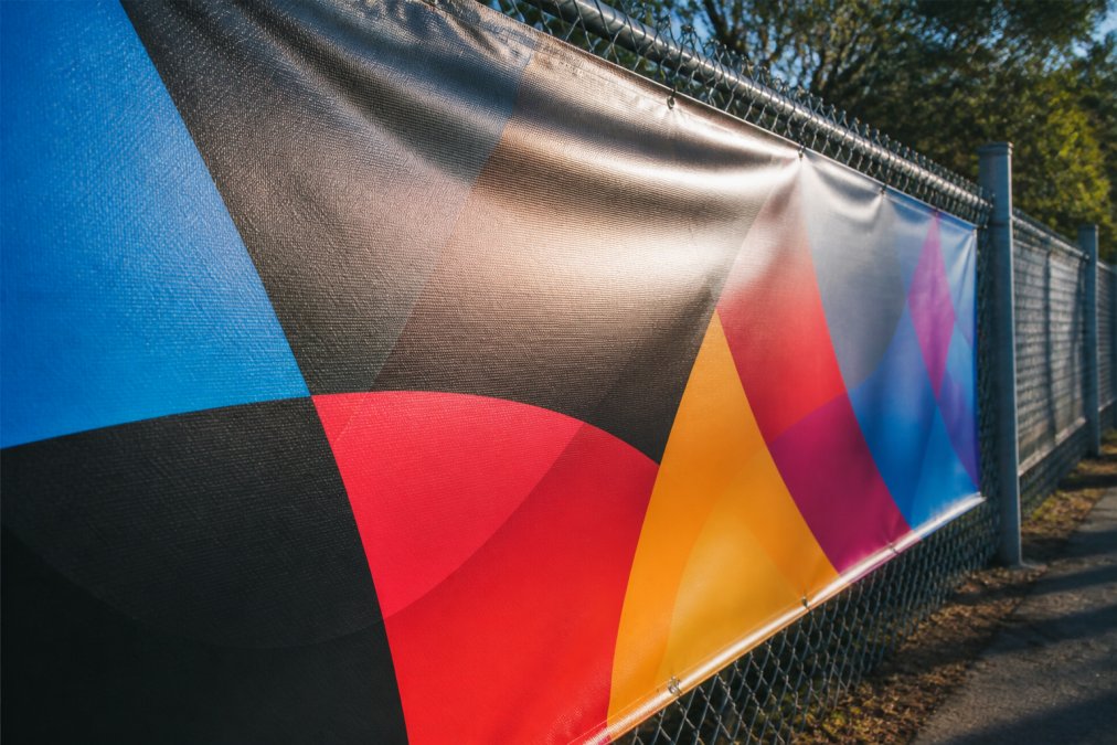 A close-up photograph of a large outdoor vinyl banner attached to a chain-link fence, showing vibrant printed colors and a slight sheen from the eco solvent ink, with afternoon sunlight hitting the surface to highlight weather resistance