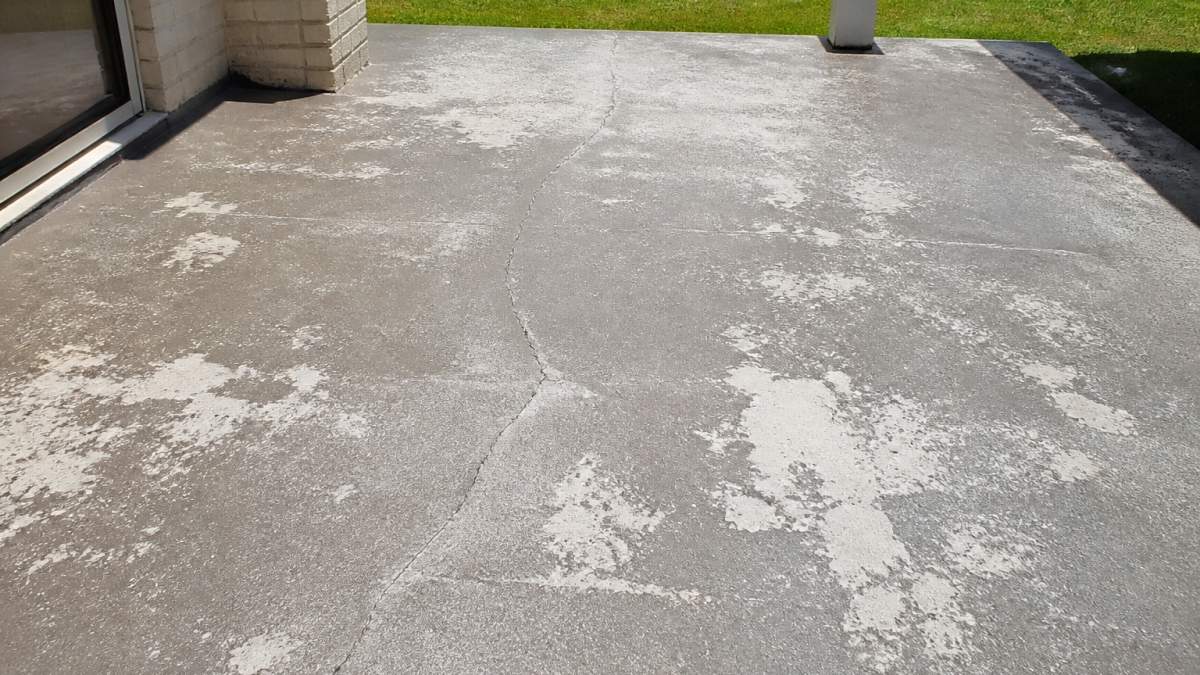 A weathered concrete patio with visible hairline cracks, faded grey surface and chalky white patches where old coating has worn through, photographed in harsh midday sunlight