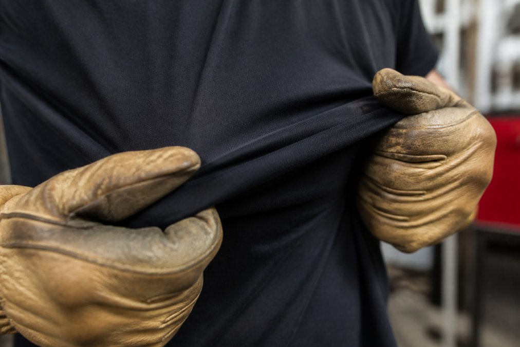 Close-up of a firefighter's hands in worn leather work gloves gripping the fabric of a black premium t-shirt, showing the tight weave and texture of the material under natural station lighting