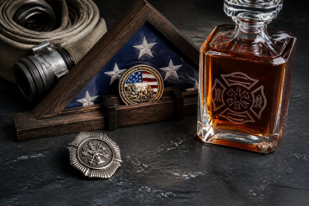 Flat lay of firefighter memorabilia including a custom engraved whiskey decanter with fire department badge design, a challenge coin displayed on a rustic wooden flag holder, and a polished firefighter badge, arranged on a dark slate surface with soft overhead lighting