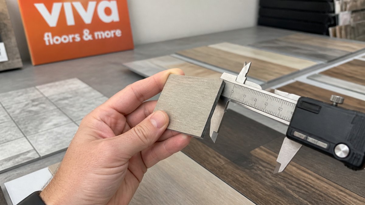 Close-up of a hand holding a small square flooring sample next to a caliper or ruler, with several LVP, laminate, and tile samples arranged on a showroom table in the background