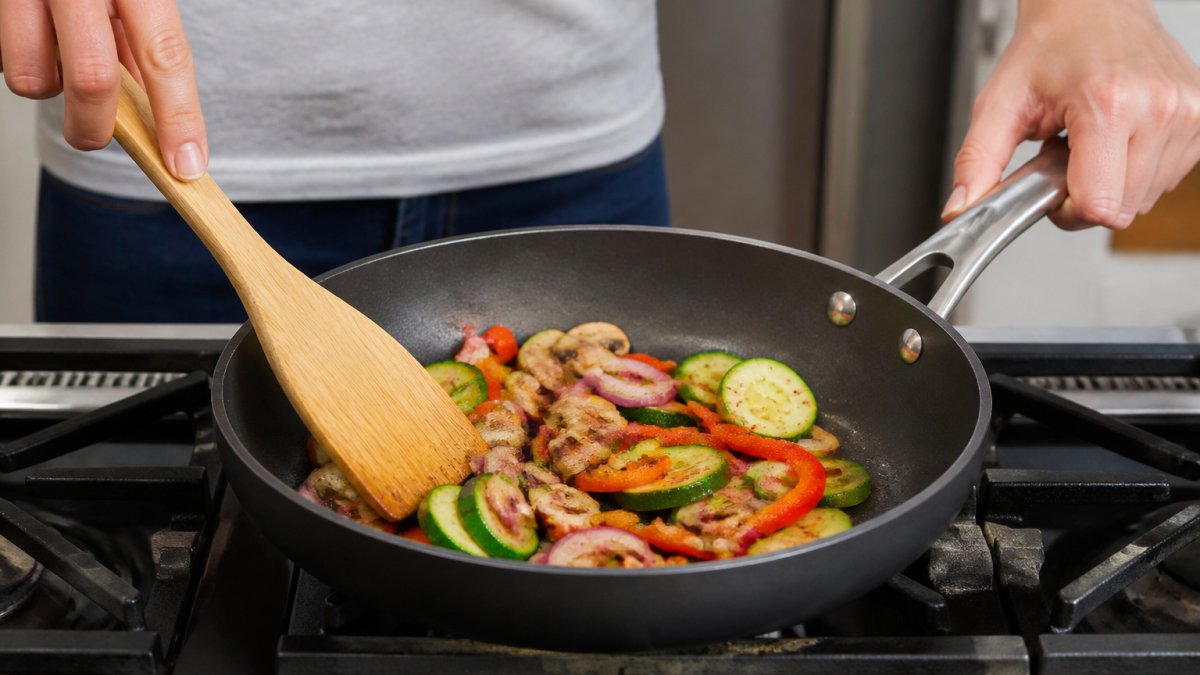 Close-up of hands preparing food in a hard-anodized fry pan on a stovetop, showing sautéed vegetables with the nonstick surface in action