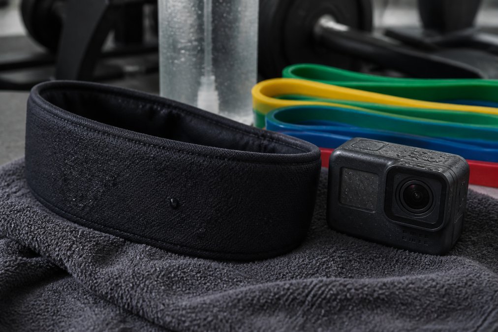 A close-up of a sweat-proof Bluetooth headband and a mini action camera resting on a gym towel, with a water bottle and resistance bands in the background, showing their durable design for intense workouts