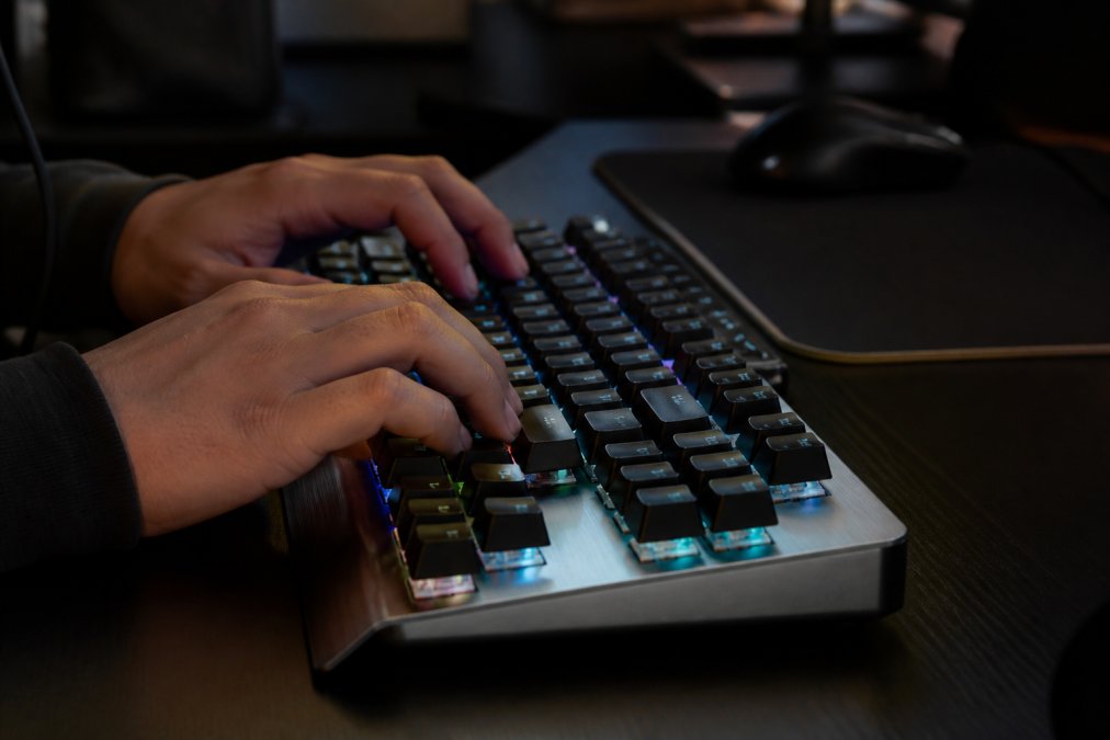 Close-up of hands typing rapidly on a mechanical gaming keyboard with a visible brushed aluminum chassis and textured PBT keycaps, illuminated by subtle RGB glow in a dimly lit gaming room