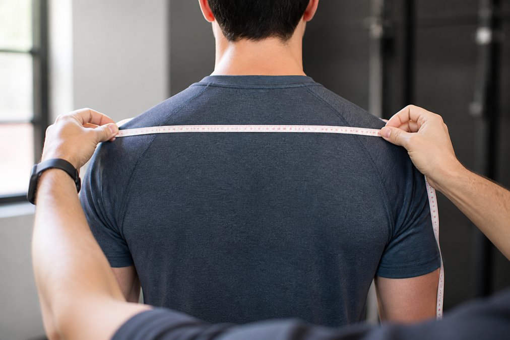Close-up of person measuring across the back of shoulders with a soft measuring tape, showing proper measurement technique from shoulder seam to shoulder seam, with clear shoulder bone reference points