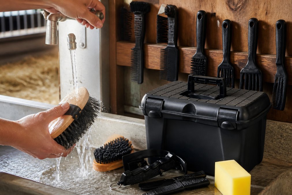 Close-up of hands with water running over stainless steel grooming brushes in a barn sink, demonstrating proper rinsing technique with natural barn lighting and organized tool drying rack in background