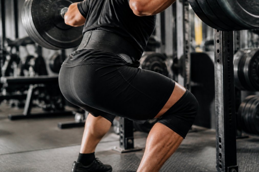 Close-up of an athlete performing a heavy barbell squat in a gym, showing reinforced seams and supportive waistband staying firmly in place as the lifter descends, with focus on the leg and hip area under bright gym lighting