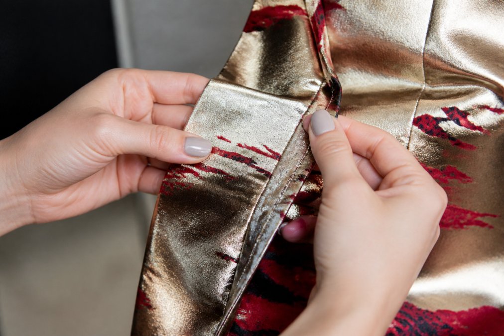 Close-up of hands examining seams and stitching on a formal dress fabric, showing detailed thread work with tight, even stitches and reinforced seam tension on metallic jacquard material