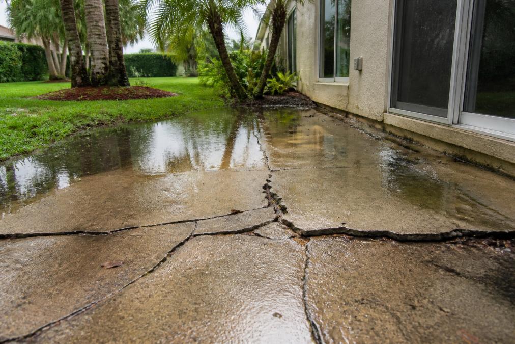 A cracked and uneven concrete patio in a lush Florida backyard after heavy rain, with standing water pooling near the foundation and surrounding palm trees
