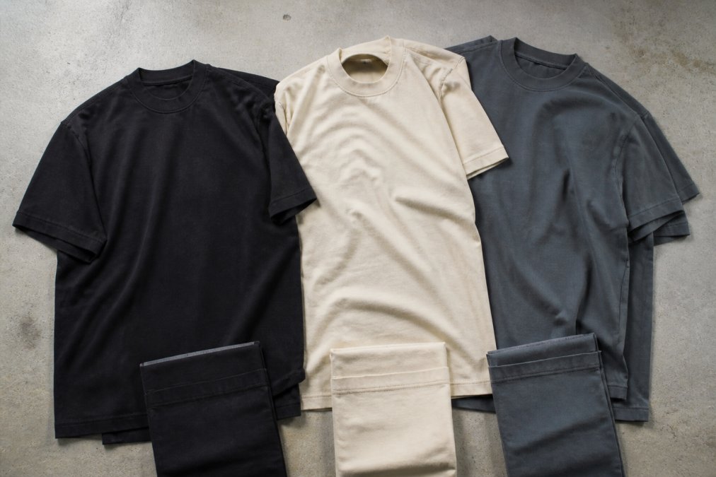 Flat lay of multiple heavyweight t-shirts in black, cream, and slate colorways arranged to showcase fabric texture, fit differences, and construction details with natural overhead lighting