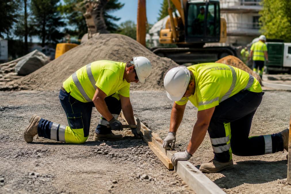 Construction workers on a busy site wearing high-visibility pants with reflective tape clearly visible, kneeling and working on ground-level tasks during daylight hours with safety helmets