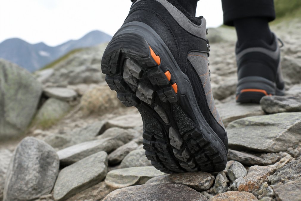 Close-up of a hiking boot sole showing aggressive multi-directional lug pattern with deep tread on rocky, uneven mountain terrain with scattered sharp stones