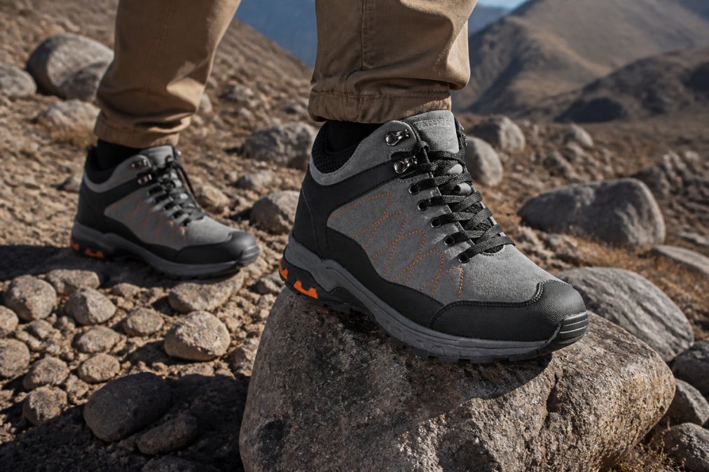 Hiker's feet wearing durable hiking boots standing on rocky, uneven trail with scattered boulders and rough mountain terrain, sunlight casting shadows