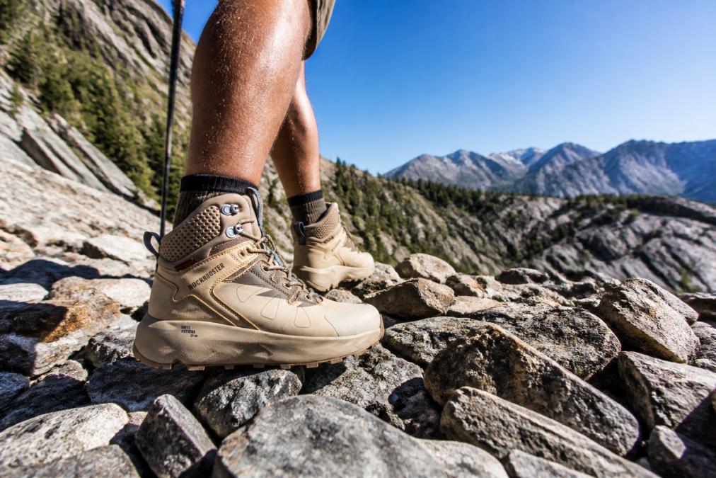 Hiker in sturdy hiking boots with aggressive tread pattern carefully navigating across a rocky scree field with sharp stones and boulders visible, bright daylight illuminating the challenging terrain