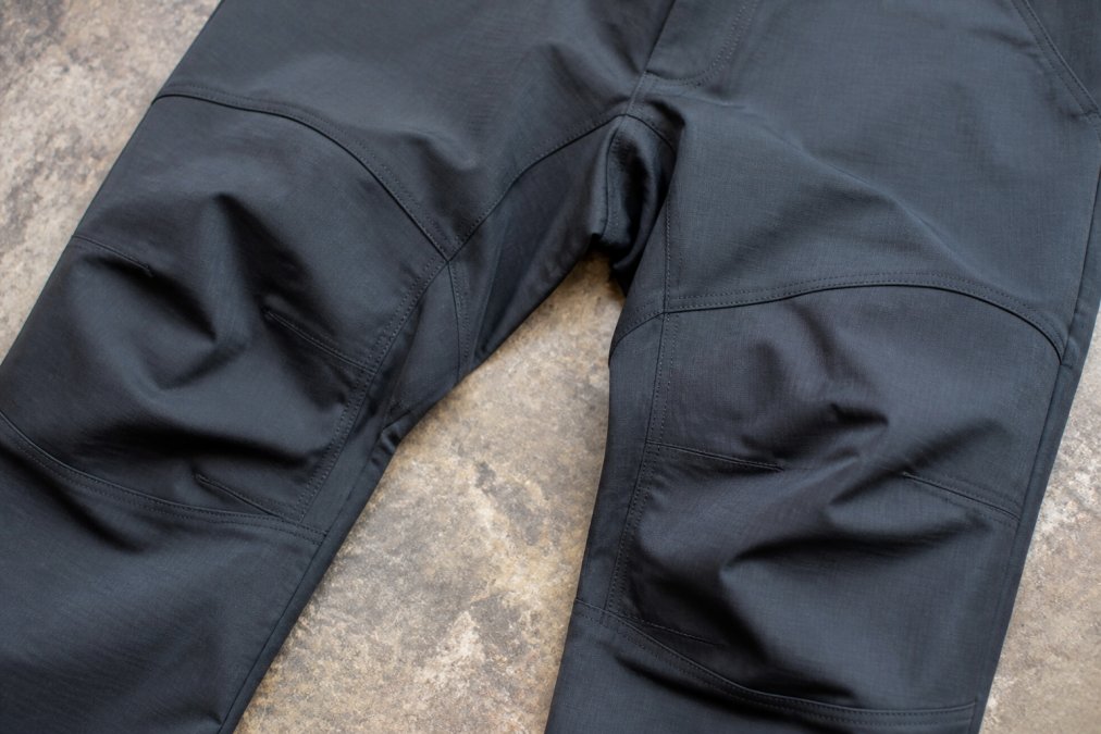 Close-up detail shot of articulated knees showing the pre-bent construction, gusseted crotch reinforcement, and four-way stretch fabric weave on technical hiking pants laid flat with visible stitching and reinforced seams
