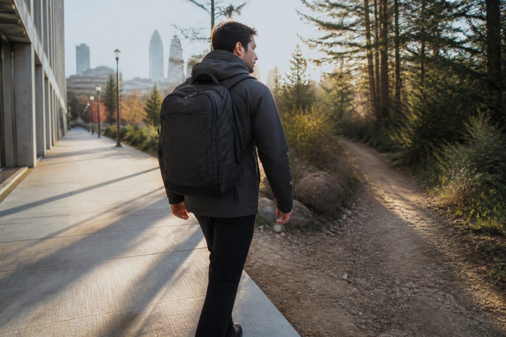 A man in a matte charcoal technical jacket and commuter backpack walking from a concrete city sidewalk onto a dirt trail at the edge of a park, morning light casting long shadows