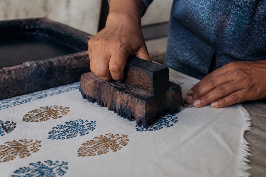 Close-up of artisan hands pressing a carved wooden block onto white cotton fabric, natural dye transferring in vibrant indigo and earth tones, showing the precise hand-stamping motion with careful detail