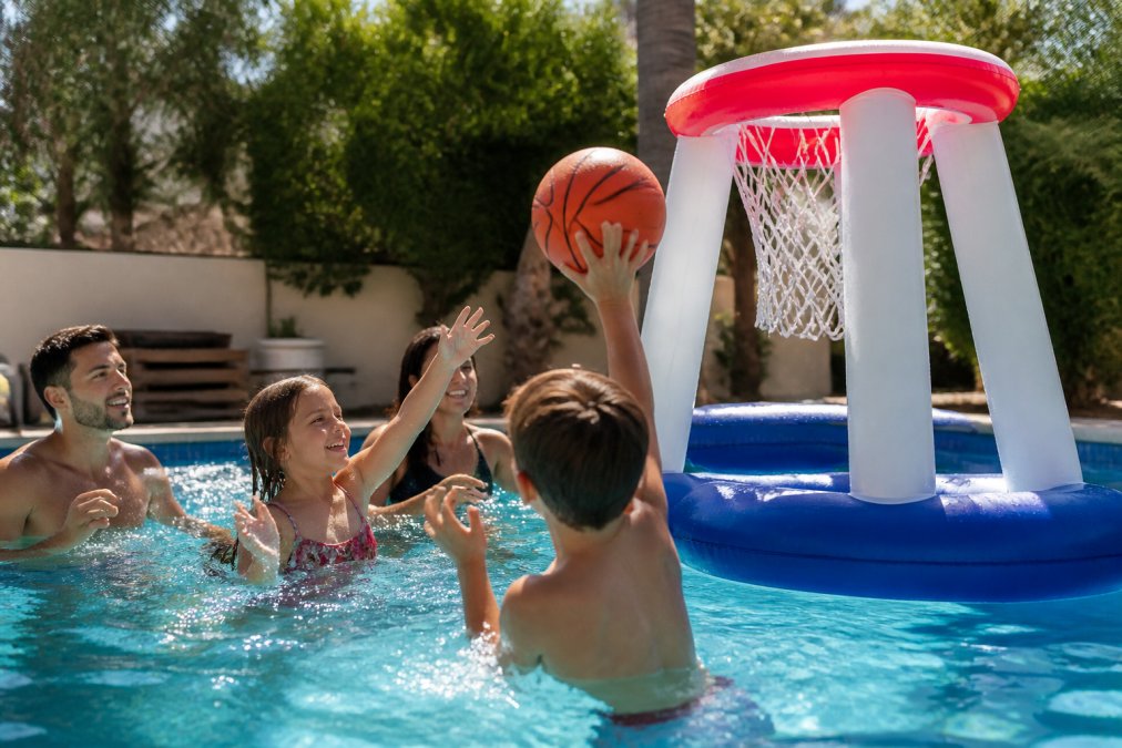 A family playing with a large inflatable basketball hoop in a sunny backyard swimming pool, the hoop floating on the water with children laughing and reaching for the ball, bright summer daylight