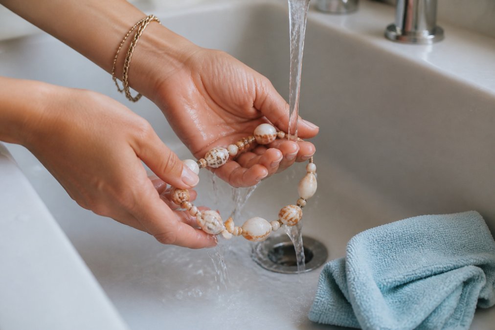 Hands gently rinsing a shell necklace under fresh running water with a soft microfiber cloth nearby on a white surface, showing proper jewelry cleaning technique in natural light