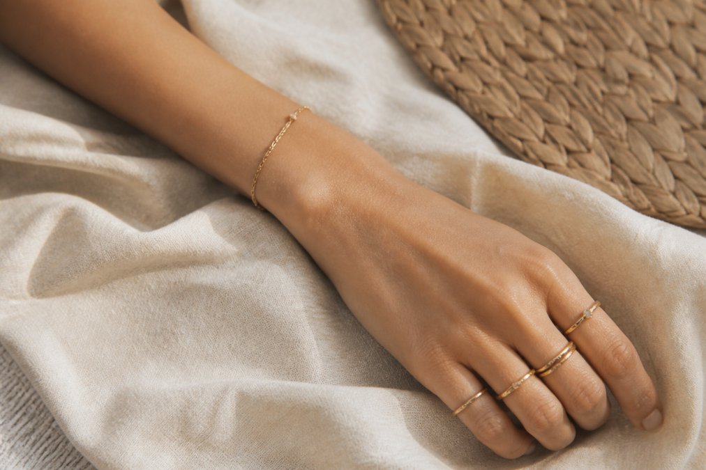 A woman's wrist and hand wearing a stack of delicate gold rings and a thin bracelet, styled against a neutral linen background in warm natural light