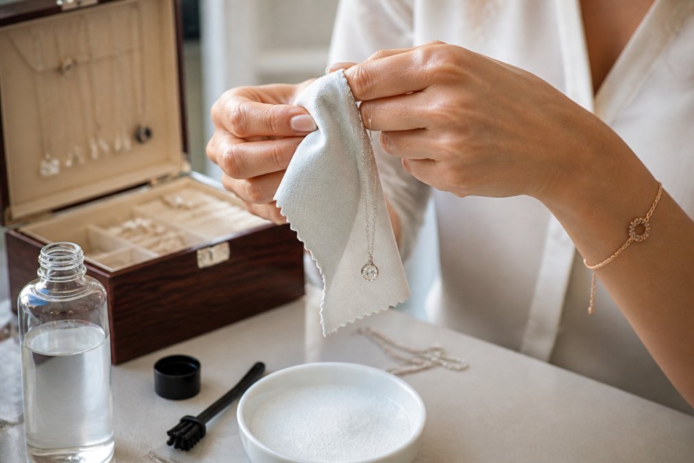 Hands gently cleaning a delicate sterling silver necklace with a soft microfiber cloth and mild polishing solution, with a wooden jewelry box and fabric-lined storage compartments visible in soft background lighting