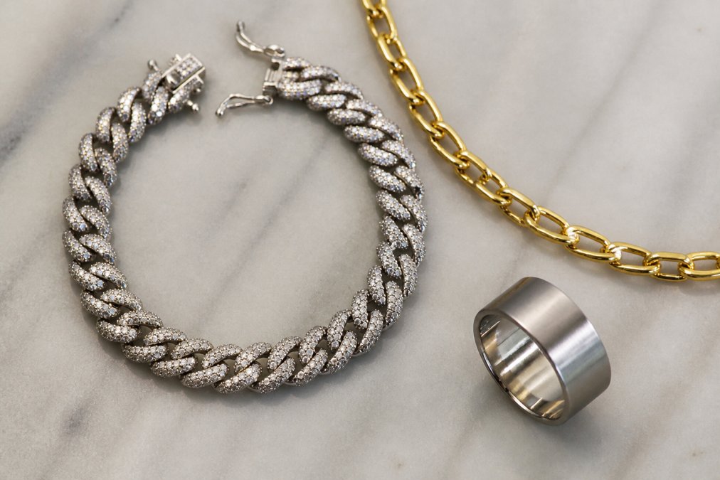 Close-up of three different jewelry pieces arranged on a neutral surface showing distinct finishes: a polished sterling silver bracelet with mirror-like shine, a warm gold-plated necklace with rich luster, and a brushed stainless steel ring with matte finish, displaying the textural differences between each metal type