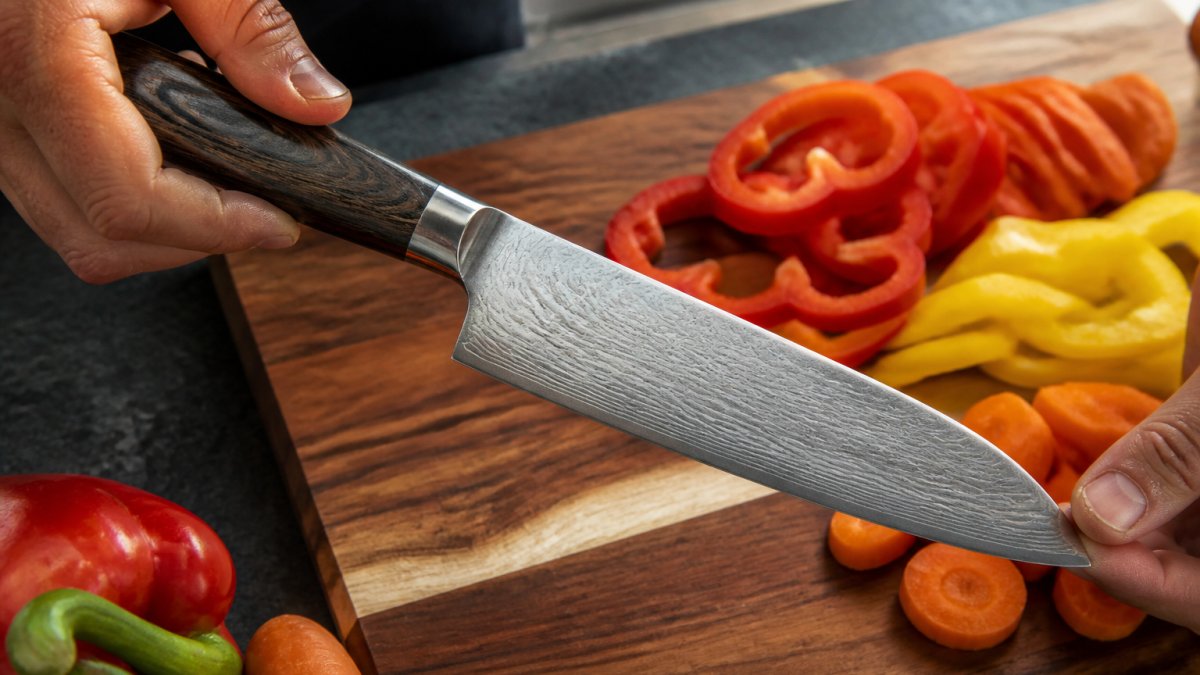 Close-up of hands holding a Damascus steel kitchen knife, showing the layered wavy blade pattern and dark wood Pakkawood handle, resting on a wooden cutting board with sliced bell peppers and carrots nearby in natural kitchen light