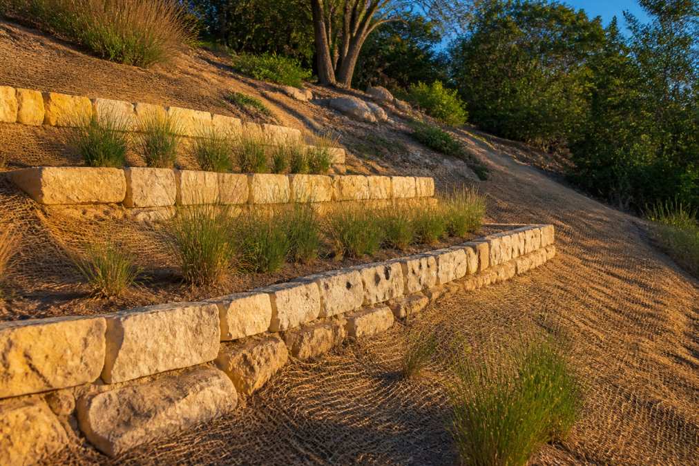 Hillside slope with limestone block retaining walls creating level terraces, native grasses planted on each terrace, and erosion control matting visible along slope contours in afternoon sunlight