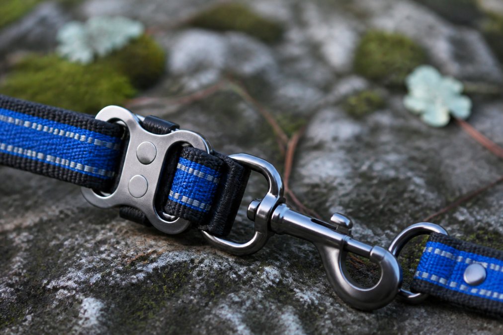 Close-up detail shot of heavy-duty dog leash hardware with swivel snap connection, reinforced stitching with visible reflective thread material, and metal reinforcement plates in natural daylight