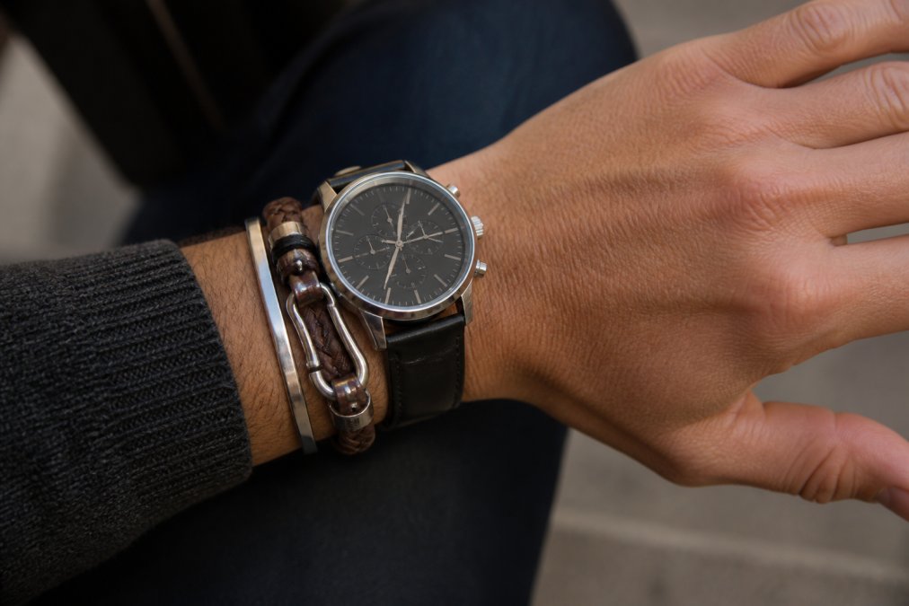 Man's wrist displaying a leather bracelet layered with a watch and stainless steel hardware, casual setting with natural lighting showing how the pieces work together harmoniously