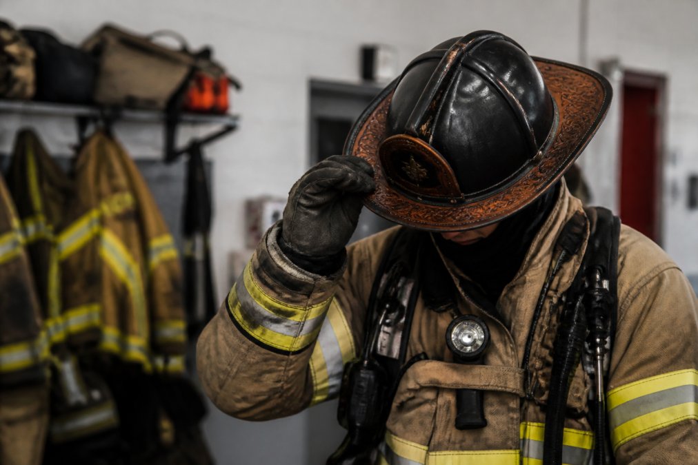 Firefighter in full turnout gear wearing a helmet with custom leather brim featuring intricate scroll work, photographed in a fire station setting with equipment in the background