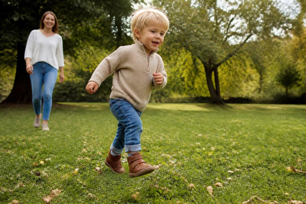 A young child wearing leather ankle boots running across a grassy park, parent smiling in the background, natural daylight, candid active play moment