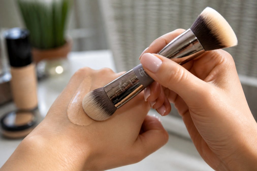 Hands holding dual-ended makeup brush demonstrating foundation application technique, with close-up showing ultra-plush bristles against skin in natural light