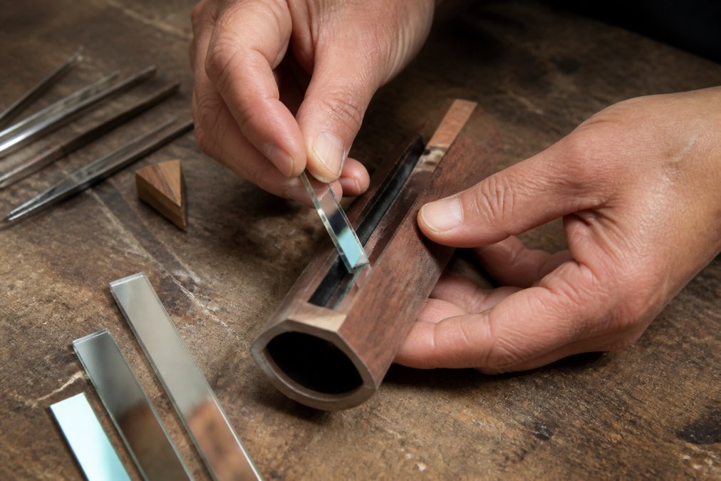 Close-up of hands carefully placing a first surface mirror strip into a wooden kaleidoscope body on a workbench, with small tools and mirror pieces nearby