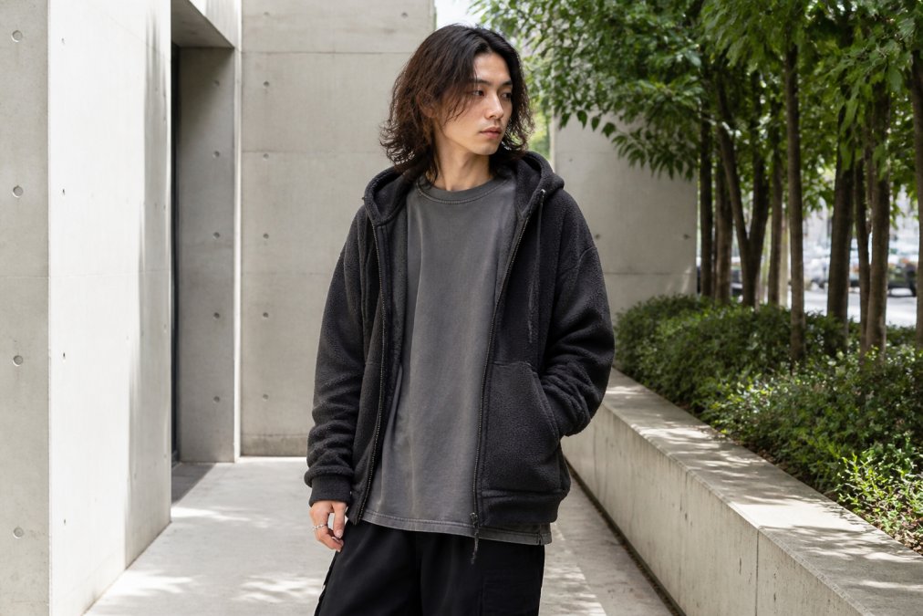 A young adult wearing an oversized vintage-washed tee layered under an open boxy fleece hoodie, standing in a bright urban setting with neutral concrete and greenery, relaxed posture, soft daylight
