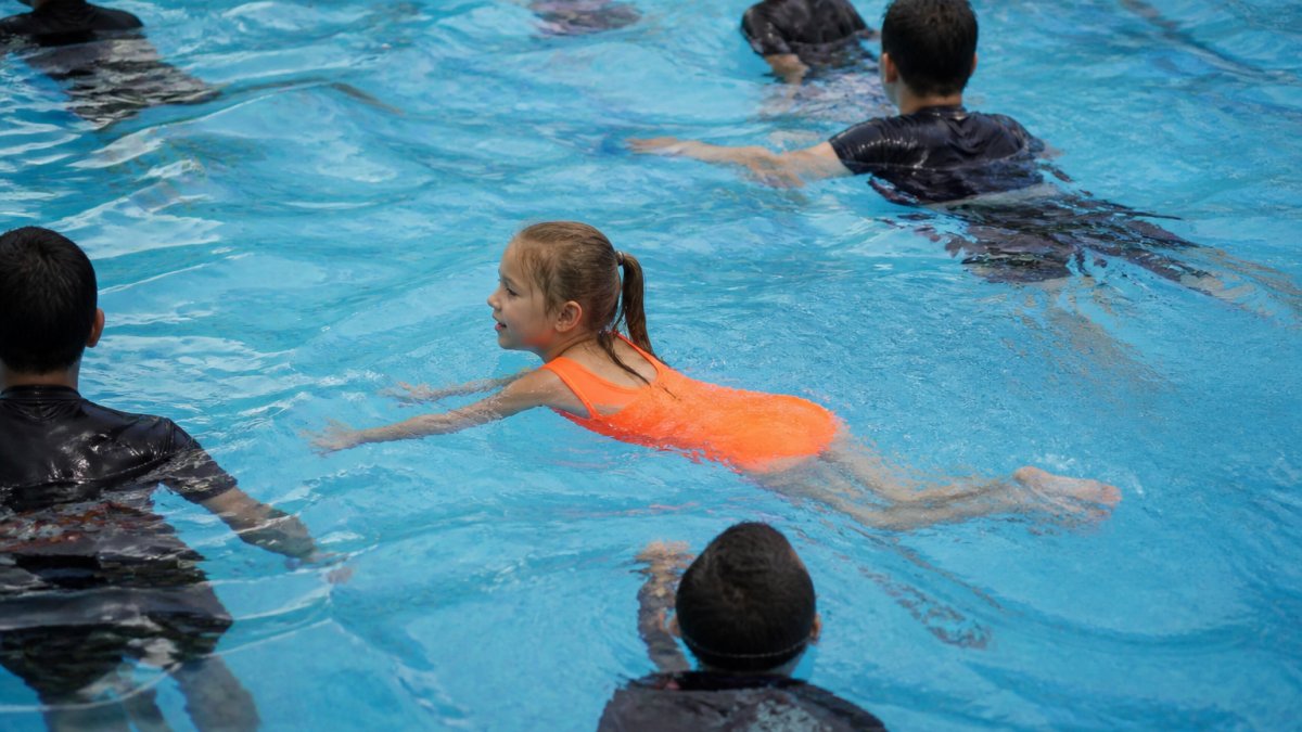 Child wearing bright neon orange one-piece swimsuit swimming in crowded pool with other swimmers in darker colors blending into blue water, easily visible against the background