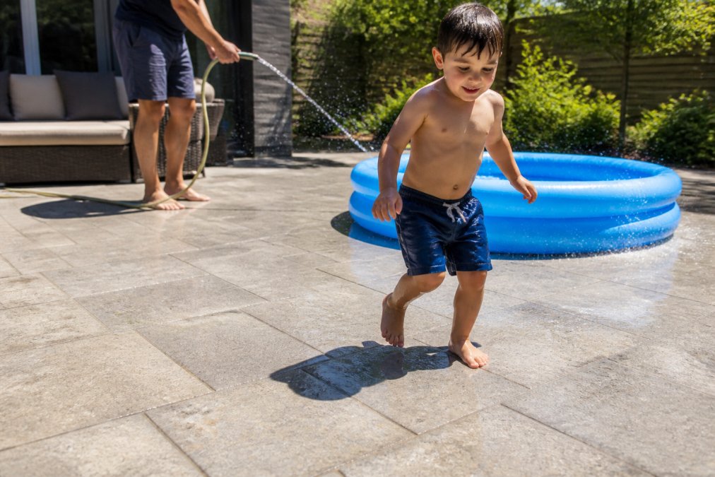 A barefoot child stepping onto a sunlit paved patio beside a wading pool, with a parent nearby holding a hose, showing the surface in bright summer light