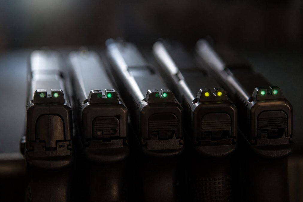 Close-up of multiple pistol sights mounted on pistol slides displayed in low-light conditions with tritium illumination glowing visibly against dark background