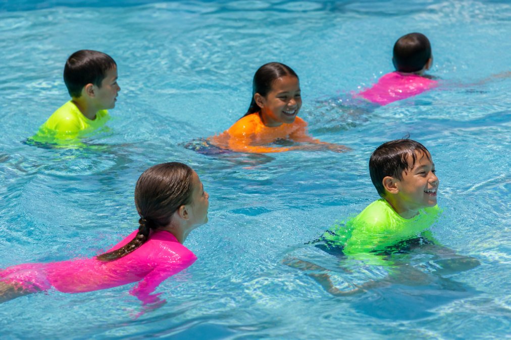 Group of active children wearing bright neon rash guards swimming and playing together in a clear pool, demonstrating comfortable movement and high visibility in water