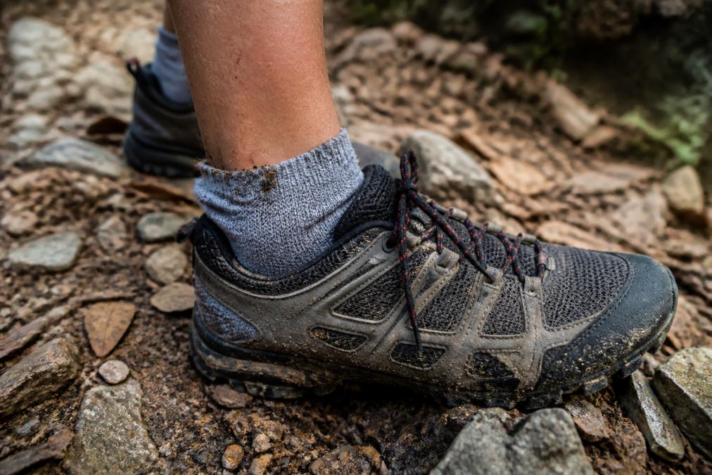 Close-up of a runner's foot in a trail running shoe on rocky, debris-filled terrain, showing the sock cuff sitting at the shoe collar with visible terrain