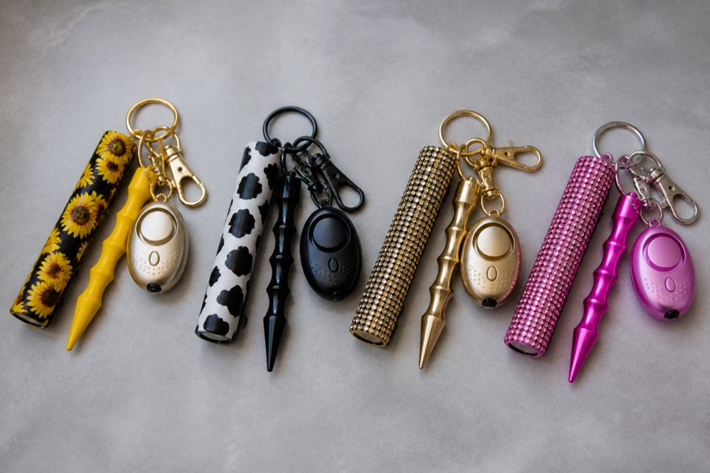 Close-up of multiple colorful safety keychains with different designs including sunflower, cow print, and rhinestone patterns arranged on neutral background, showing compact size and attachment hardware