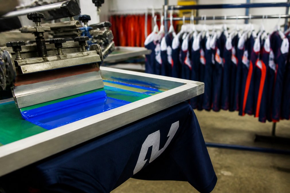 Close-up of a screen printing press applying vibrant ink onto a youth sports jersey in a print shop, with a rack of finished team uniforms in the background